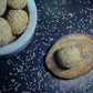 A close-up image of traditional Indian sweets, til (sesame) laddus, some roasted and displayed on a wooden surface with a bowl in the background, scattered with sesame seeds.